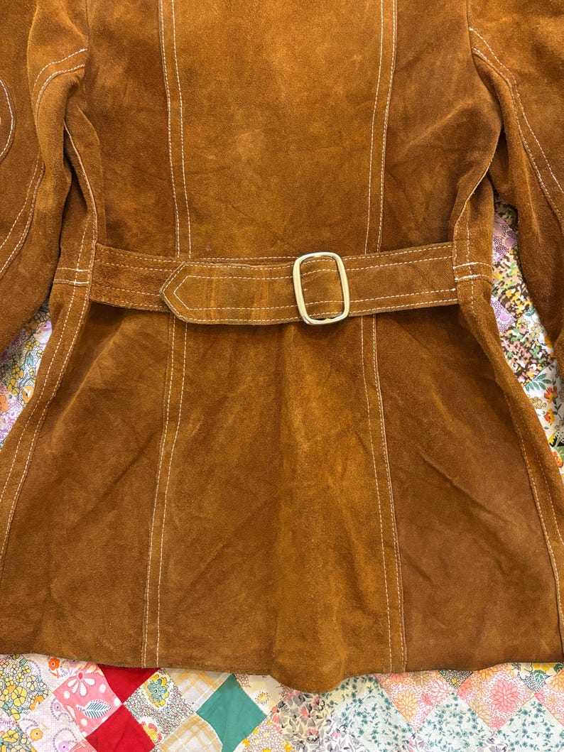 Puede incluir: Chaqueta de ante marr&oacute;n con cintur&oacute;n a juego y hebilla dorada. La chaqueta presenta costuras blancas a lo largo de las costuras y un corte entallado. La chaqueta es de estilo vintage.