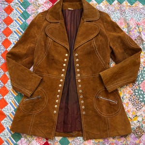 Puede incluir: Chaqueta vintage de ante marr&oacute;n con cuello con solapas, m&uacute;ltiples botones y dos bolsillos delanteros con cremalleras. La chaqueta tiene detalles de costuras blancas y un forro interior marr&oacute;n oscuro. Sobre un edred&oacute;n patchwork colorido.