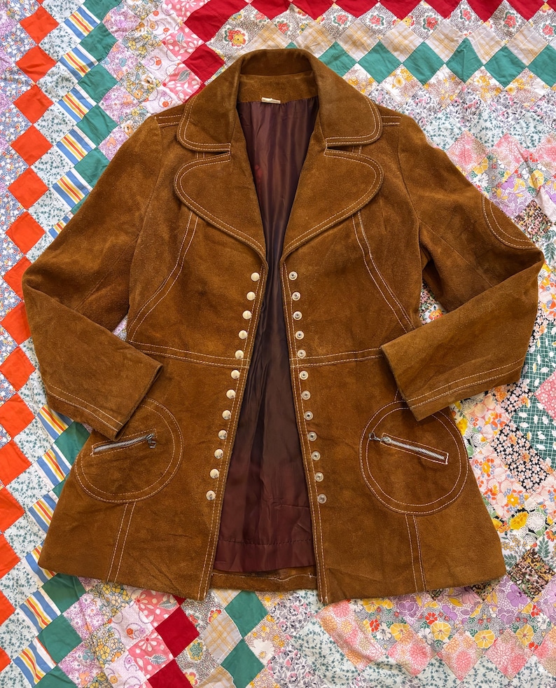 Puede incluir: Chaqueta vintage de ante marr&oacute;n con cuello con solapas, m&uacute;ltiples botones y dos bolsillos delanteros con cremalleras. La chaqueta tiene detalles de costuras blancas y un forro interior marr&oacute;n oscuro. Sobre un edred&oacute;n patchwork colorido.