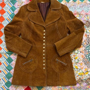 Puede incluir: Chaqueta vintage de ante marr&oacute;n con cuello con solapas, cierre de botones y dos bolsillos delanteros con cremalleras. La chaqueta presenta costuras blancas y un corte entallado, evocando una est&eacute;tica retro.
