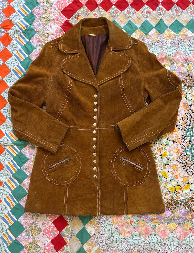 Puede incluir: Chaqueta vintage de ante marr&oacute;n con cuello con solapas, cierre de botones y dos bolsillos delanteros con cremalleras. La chaqueta presenta costuras blancas y un corte entallado, evocando una est&eacute;tica retro.