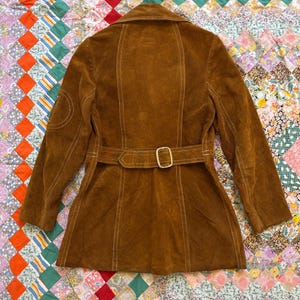 Puede incluir: Chaqueta de ante marr&oacute;n con cintur&oacute;n y cuello. La chaqueta tiene costuras blancas y un parche en la manga izquierda. El cintur&oacute;n tiene una hebilla dorada. La chaqueta se muestra sobre un edred&oacute;n patchwork colorido.