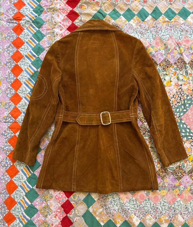 Puede incluir: Chaqueta de ante marr&oacute;n con cintur&oacute;n y cuello. La chaqueta tiene costuras blancas y un parche en la manga izquierda. El cintur&oacute;n tiene una hebilla dorada. La chaqueta se muestra sobre un edred&oacute;n patchwork colorido.