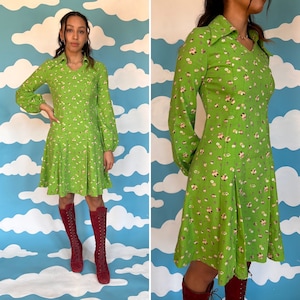 Könnte beinhalten: Ein grünes Kleid mit Blumenmuster und Kragen. Das Kleid hat lange Ärmel mit einem gerafften Ärmelabschluss und einen ausgestellten Rock. Das Model trägt rote Stiefel.