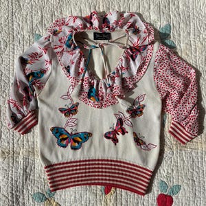 May include: Cream-coloured sweater with a red and white striped bottom hem and cuffs. The sweater features a ruffled collar and sleeves with a floral pattern. Colourful butterfly appliqués are on the front.
