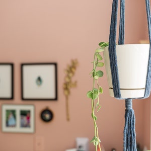 May include: A white ceramic plant pot with a trailing green plant hangs in a blue macrame plant hanger. The plant hanger is suspended in front of a pink wall with framed artwork.