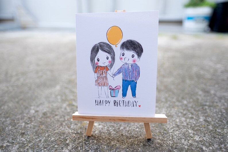 Puede incluir: Una tarjeta blanca con una ilustraci&oacute;n dibujada a mano de una pareja que se toma de la mano con un globo amarillo y una caja de regalo. El texto "Happy Birthday" est&aacute; escrito debajo de la ilustraci&oacute;n.