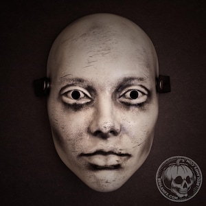 Op de afbeelding: Een bleek, mensachtig gezichtsmasker met een verweerde, versleten afwerking met donkere schaduwen rond de ogen en mond. Het masker heeft twee ronde oogopeningen en zwarte zijbanden met metalen klinknagels. Een rond logo met een schedel is zichtbaar.