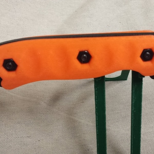 May include: Close-up of an orange and black knife handle with four black screws. The blade is black and has the text "BK-16" on it.
