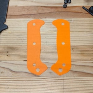 May include: Two orange knife scales with holes for mounting hardware. The scales are on a wooden surface.