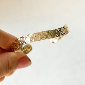 May include: A gold-colored cuff bracelet with a hammered texture. The bracelet has a slightly curved shape and a textured surface, reflecting light. The jewelry is a simple, elegant accessory.