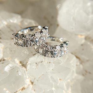 May include: Two silver hoop earrings with oval-shaped clear gemstones. The earrings are on a white background.