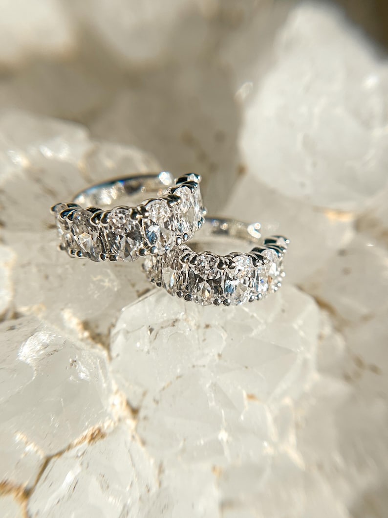 May include: Two silver hoop earrings with oval-shaped clear gemstones. The earrings are on a white background.