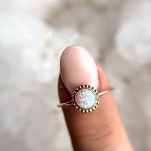 May include: A delicate gold ring with a white opal gemstone set in a beaded bezel. The ring is on a finger.