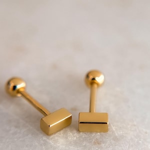 May include: Two gold-colored stud earrings. Each earring has a small, round ball at the top and a rectangular bar at the bottom. The earrings are set against a light-colored, marble-like surface.