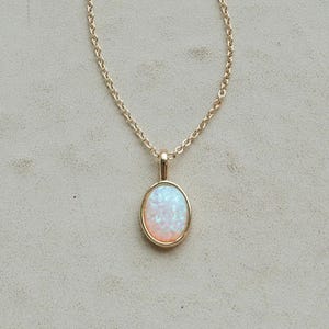 May include: A gold-colored necklace with a round pendant. The pendant features a white and light blue opal stone set in a gold bezel. The chain is delicate and also gold-colored, creating a simple and elegant design.