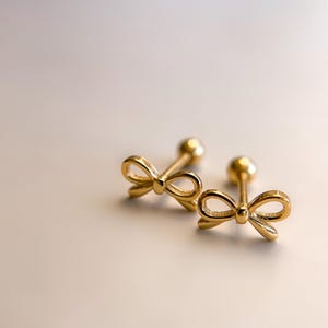 以下が含まれることがあります： 2つの金色のリボン型スタッドピアス。ピアスには小さくて丸いポストと繊細なリボンデザインがあります。ピアスは明るい色の表面に表示されています。