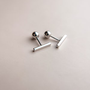May include: Two silver cufflinks with a ball-shaped head and a rectangular bar. The cufflinks are displayed on a light pink surface. The design is simple and modern, suitable for formal wear.
