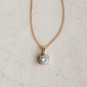 Op de afbeelding: Een goudkleurige ketting met een grote, ronde, heldere edelsteen hanger. De hanger is gezet in een vier-klauw zetting en hangt aan een fijne ketting. De achtergrond is een neutrale, gebroken witte kleur.