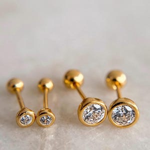 May include: Four gold-tone stud earrings with round, clear, faceted stones set in a bezel setting. Each earring features a thin post and a smooth, spherical back closure. The set includes two smaller earrings and two larger earrings, all matching.