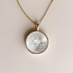 Könnte beinhalten: Eine goldene Kettenkette mit einem runden Anhänger, der ein weißes, graviertes Design eines Halbmondes und Sonnenstrahlen zeigt.