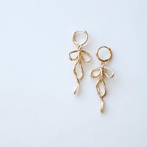 Puede incluir: Un par de pendientes colgantes dorados. Cada pendiente presenta un pequeño aro en la parte superior, conectado a una forma de lazo estilizada, con un diseño ondulado, alargado y fluido. Los pendientes están sobre un fondo blanco.