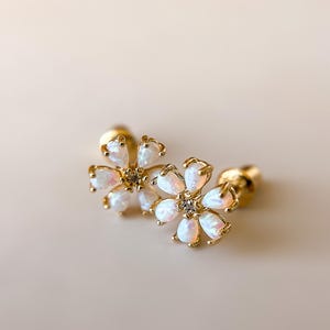 Puede incluir: Un par de pendientes de oro en forma de flor con piedras de ópalo blanco y un pequeño cristal transparente en el centro de cada flor.