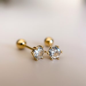 May include: A pair of gold stud earrings with clear round cubic zirconia stones. The earrings have a simple, classic design.