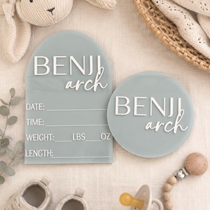 Puede incluir: Dos letreros de acrílico verde salvia con letras blancas en 3D que dicen BENJI arch. El letrero arqueado incluye campos para fecha, hora, peso en lbs y oz, y longitud. El letrero circular presenta el mismo nombre. Ambos tienen acabado mate.