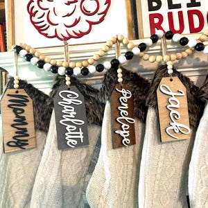 May include: Christmas stockings hanging from a mantel, each with a name tag. The stockings are white cable knit with faux fur trim. A wooden bead garland hangs above. A red Santa face and a sign that says "Don't Blame Rudolph" are in the background.
