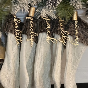 May include: Five white cable knit Christmas stockings hang from a decorated mantle. Each stocking has a faux fur cuff and a gold name tag. The names on the tags are Charlotte, Penelope, Jack, Kaddy, and Tommy. The mantle is decorated with pine and frosted pinecones.
