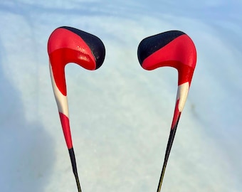 May include: A pair of black, red, and white wireless earbuds with a long, thin wire connecting them. The earbuds are held in a hand against a white background.