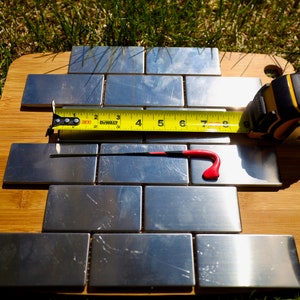 May include: A close-up of a set of stainless steel tiles on a wooden cutting board. A yellow measuring tape is placed across the tiles, with the numbers 2 through 9 visible. A red tool is also visible on the tiles.