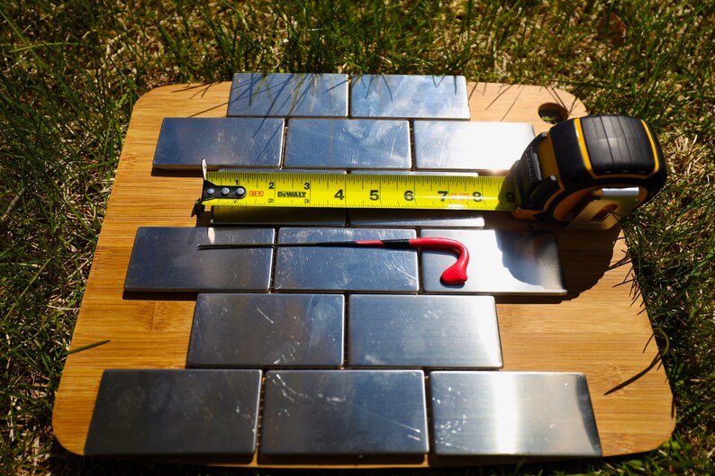 May include: A close-up of a set of stainless steel tiles on a wooden cutting board. A yellow measuring tape is placed across the tiles, with the numbers 2 through 9 visible. A red tool is also visible on the tiles.