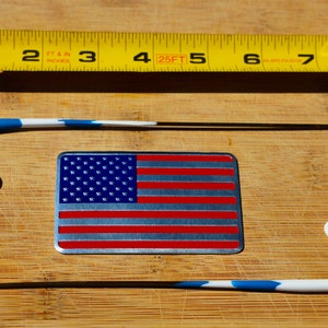 May include: Two white and blue earbud hooks with a metal American flag sticker on a wooden surface. The sticker has red and white stripes and blue stars.