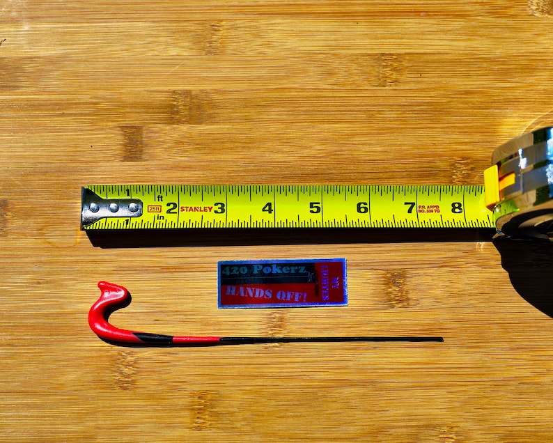 May include: A yellow measuring tape with black markings and a silver case. The tape reads "Stanley" and has markings for inches and feet. A red and black hook is also pictured. A sticker with the text "420 Pokerz Hands Off!" is visible.