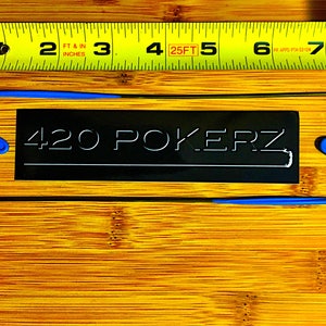 May include: A black rectangular sign with the text "420 POKERZ" in white lettering. Two blue and black metal hooks are on either side of the sign. A yellow measuring tape is in the background.