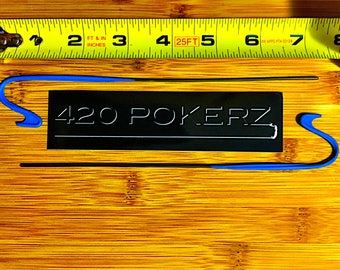 May include: A black rectangular sign with the text "420 POKERZ" in white lettering. Two blue and black metal hooks are on either side of the sign. A yellow measuring tape is in the background.