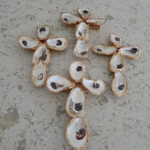 May include: Arrangement of oyster shells, each with a gold-painted edge, forming cross shapes. The shells are white with a dark brown interior, set on small wooden supports. The background is a light, speckled surface.