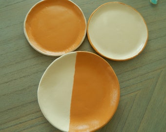 May include: Three handmade ceramic plates with a modern design. Two plates are orange with white rims, one is half orange and half white, and the third is white with an orange rim. The plates are on a wooden surface.
