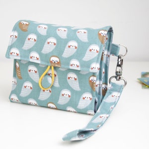 Könnte beinhalten: Eine blaugrüne Stoffbörse mit einem weißen Eulen-Druck und einem gelben Riemen. Die Börse hat einen Druckknopfverschluss und einen abnehmbaren Riemen mit einem silbernen Clip.