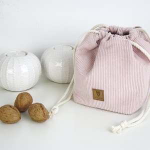 Puede incluir: Una bolsa de cord&oacute;n de pana rosa con un cord&oacute;n blanco y una etiqueta de cuero marr&oacute;n con un logotipo de coraz&oacute;n. La bolsa est&aacute; sobre una superficie blanca con dos candelabros de cer&aacute;mica blanca y tres nueces.