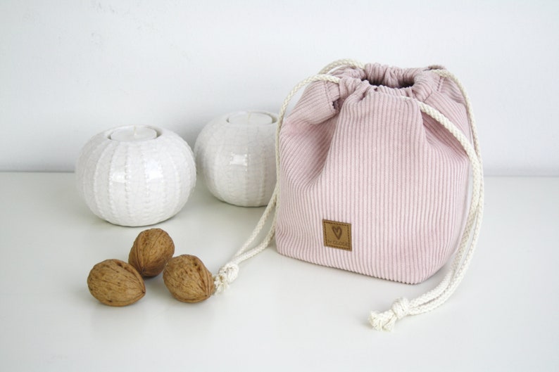 Puede incluir: Una bolsa de cord&oacute;n de pana rosa con un cord&oacute;n blanco y una etiqueta de cuero marr&oacute;n con un logotipo de coraz&oacute;n. La bolsa est&aacute; sobre una superficie blanca con dos candelabros de cer&aacute;mica blanca y tres nueces.
