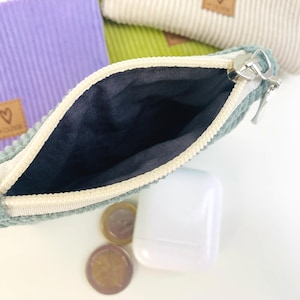 May include: A small, green corduroy zippered pouch with a white zipper and a black lining. The pouch is open and the zipper is pulled down. The pouch is sitting on a white surface with some coins and a white case.