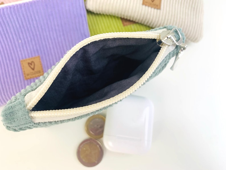 May include: A small, green corduroy zippered pouch with a white zipper and a black lining. The pouch is open and the zipper is pulled down. The pouch is sitting on a white surface with some coins and a white case.