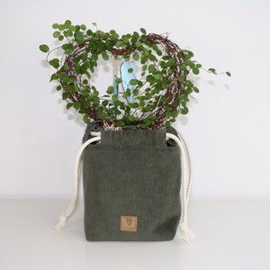 May include: A green corduroy planter with white drawstrings and a heart-shaped wreath of green foliage inside. The planter has a brown leather tag with a heart symbol.