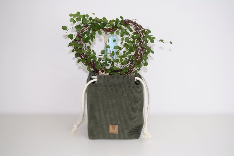 May include: A green corduroy planter with white drawstrings and a heart-shaped wreath of green foliage inside. The planter has a brown leather tag with a heart symbol.