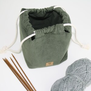 May include: A green corduroy drawstring bag with white drawstrings, a ball of grey yarn, and wooden knitting needles.