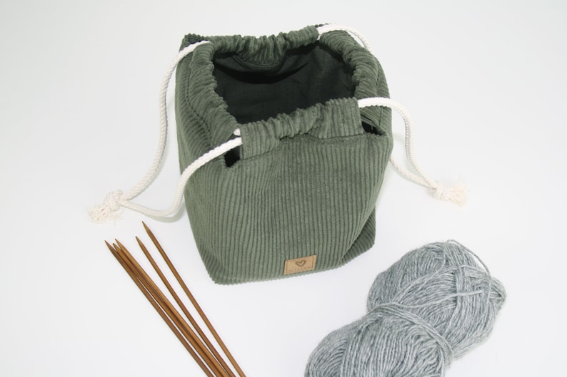 May include: A green corduroy drawstring bag with white drawstrings, a ball of grey yarn, and wooden knitting needles.