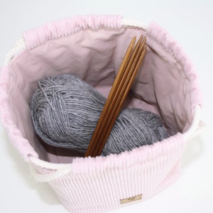 Puede incluir: Una bolsa de cord&oacute;n de pana rosa con forro blanco. La bolsa est&aacute; llena de dos ovillos de hilo gris y un juego de agujas de tejer de madera.